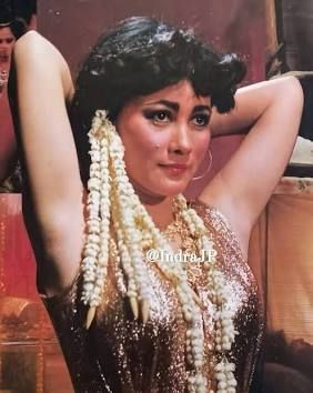 Suzanna in NYI BLORONG (1982) wearing a gold top, and her hair cascades with flowers. Her arms are raised as she gathers her hair. 