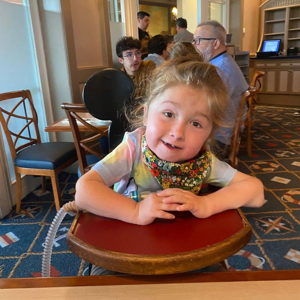 Daisy is sat in a highchair inside a restaurant at Disneyland Paris. She is smiling for the camera, wearing a tie-dye t-shirt and a floral-patterned neck scarf.