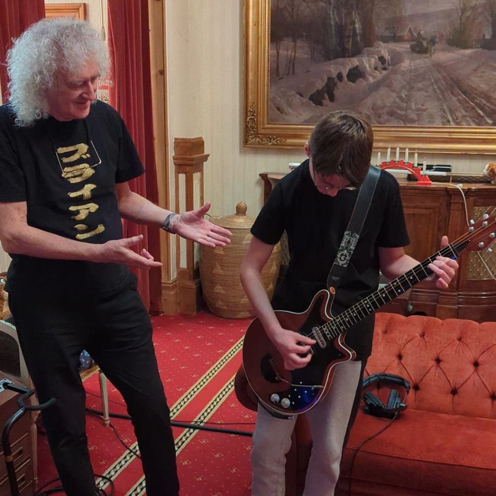 Sir Brian May and Jacob are stood in a large, carpeted living room. On the right, Jacob is playing a red guitar and concentrating on his finger positions on the strings, and he strums them. Sir Brian is stood on the left. He has both hands out gesturing towards Jacob and is smiling.