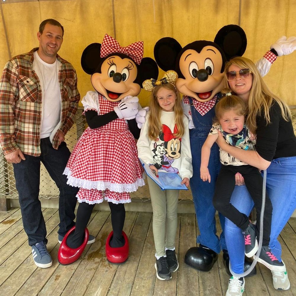 From right to left, Mum is holding Daisy, Mickey Mouse, Freya, Daisy's older sister, Minnie Mouse and Dad are all stood for a family photo together. Minnie Mouse is wearing a pink and white gingham dress and hairbow. Mickey Mouse's shirt matches this but is beneath blue dungarees. Both Freya and Daisy are wearing Mickey and Minnie shirts, and Freya also has golden ears on.