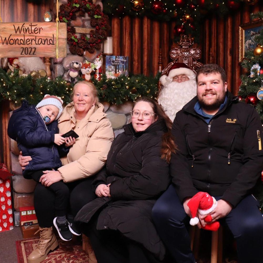Teddy and his family are sat inside a wooden log cabin at Centre Parcs’ 2022 Winter Wonderland. Behind them is Father Christmas and they’re all smiling for the camera. In the background, there are toys, presents and a Christmas tree.