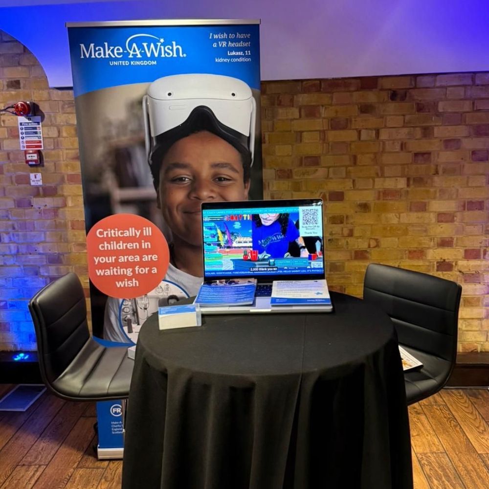 A promotional stand for Make-A-Wish UK set in front of a red and brown brick wall. A the back of the stand, there's a banner with information and a photo of a wish child wearing a white VR set on top of their head. In front of the banner, there's a black table and two tall chairs. On the table, there's a laptop with a colorful gaming show reel playing on screen.