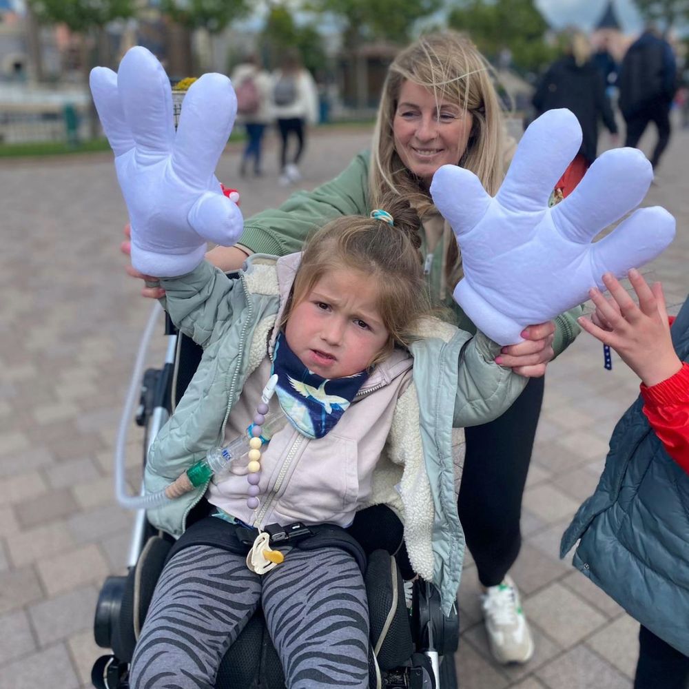 Daisy is in her wheelchair inside the Disneyland Paris Park.  Daisy's arms are raised in the air to cheer. Daisy is wearing oversized, Mickey Mouse hand gloves. Her sister Freya is stood slightly out of shot but can be seen touching one of the gloves. Mum is smiling behind Daisy.