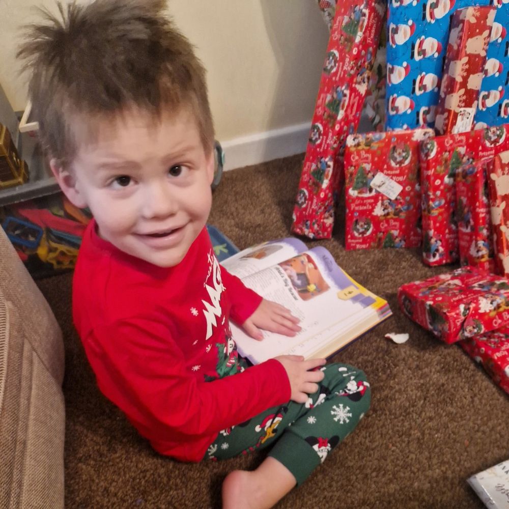 Teddy is wearing red and green Christmas pyjamas and is sat on a brown carpet. He is looking up at the camera and smiling while holding a children’s book in his hands. To the right, there are lots of Christmas presents stacked up against the wall. The wrapping paper is blue and red and has cartoon graphics of Father Christmas and Christmas trees on it.