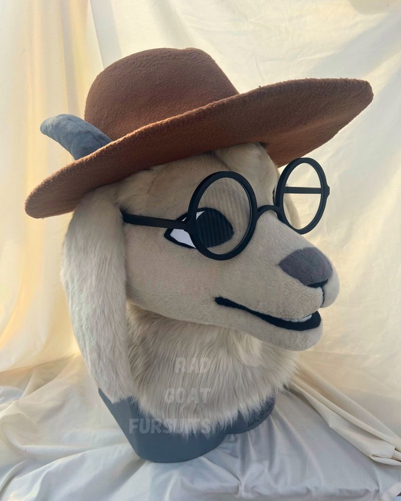 An off-white goat fursuit head with a grey spot on the nose, grey horns, and big round glasses. He is wearing a brown hat, somewhat like a detective hat. 