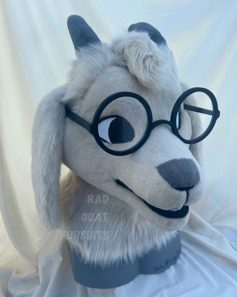 An off-white goat fursuit head with a grey spot on the nose, grey horns, and big round glasses