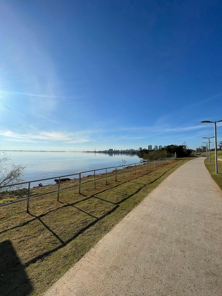 uma calçada com vista para uma lagoa, e grama ao redor. o céu está bem azul com poucas nuvens e existem alguns postes de luz acompanhando a calçada. prédios podem ser vistos a distância 