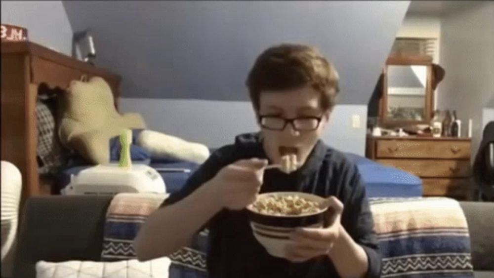 a boy with glasses is eating cereal in a bedroom