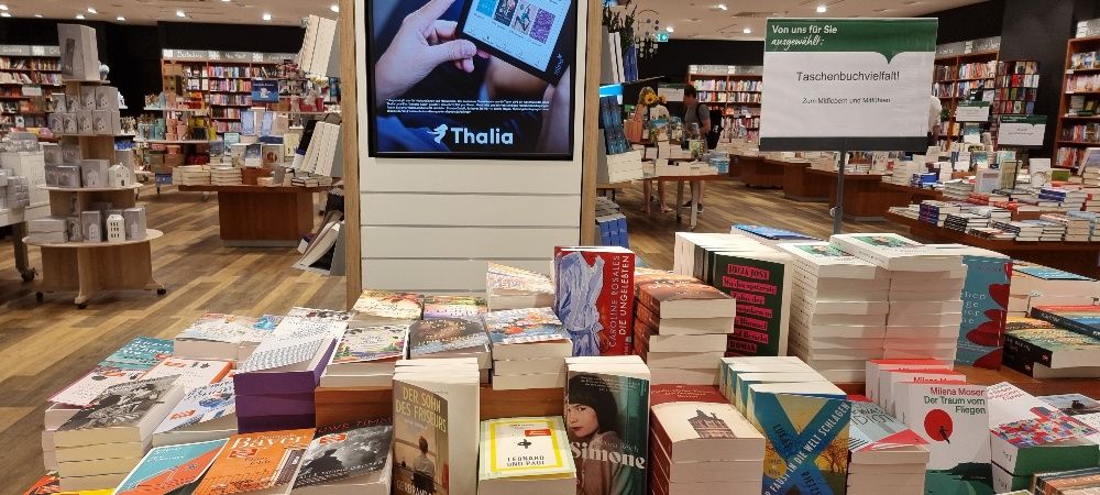 Ein Tisch mit Taschenbüchern. Darüber ein Schild: Thalia. Ganz links ein Stoß mit dem Taschenbuch "Schneeflocken wie Feuer" von Elfi Conrad. 