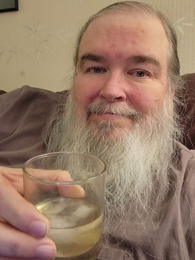 Smiling white guy, ponytailed hair, white-gray beard, gray t-shirt, on a couch, holding a glass of pale scotch whiskey on the rocks.