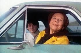 Film Harold and Maude
Maude sitzt auf dem Fahrersitz eines Autos, das sie gerade gestohlen hat und grinst in die Kamera. Harold sitzt verstört auf dem Beifahrersitzt