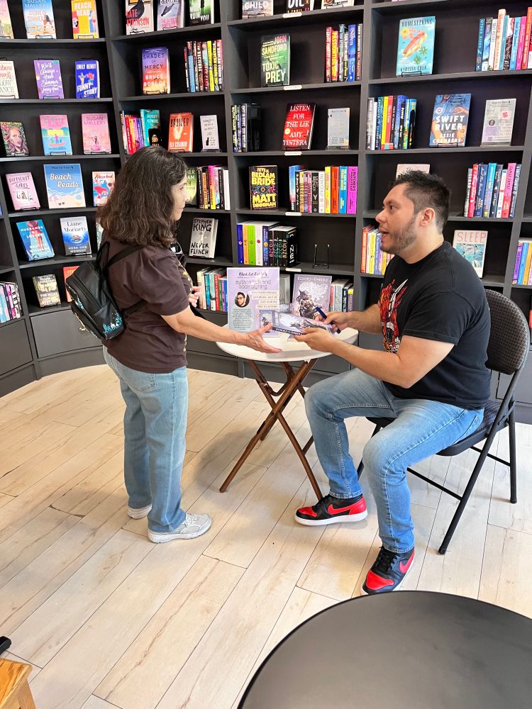 Black Cat Fables bookstore signing for Pedro Iniguez. Picture of Pedro in a StokerCon shirt and blue jeans signing his short story collection, ECHOES AND EMBERS: SPECULATIVE STORIES for a woman in jeans and a brown shirt.