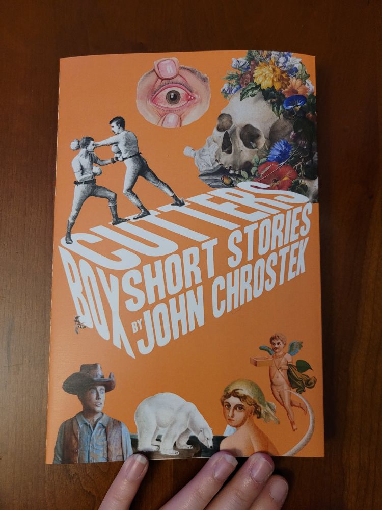 On a shiny wooden tabletop, a paperback copy of Box Cutters (short stories by John Chrostek) is centered. Four fingers touch the bottom of the cover, which is orange with white text and has assorted images on it (a polar bear, boxers boxing, a cowboy, a skull covered on flowers, a wide open eye, etc).