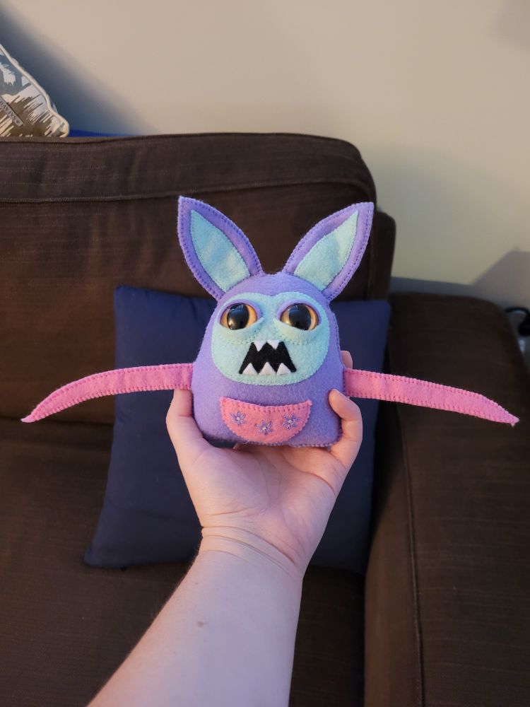 A hand is holding a hand-stitched felt creature in front of a brown couch. This is Sweet Theodore. He's a purple rabbit with a greenish blue face, large dark yellow eyes, and an open mouth filled with sharp teeth. He has two pink tentacles and a pink pouch over his belly with three purple embroidered flowers.