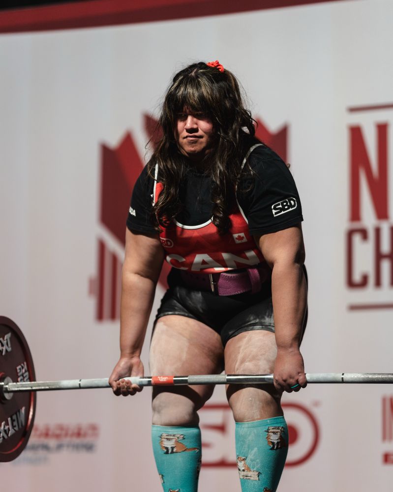 Lanie is in a powerlifting competition. She is in the middle of a convention deadlift. She is wearing a team Canada singlet and blue fox socks.