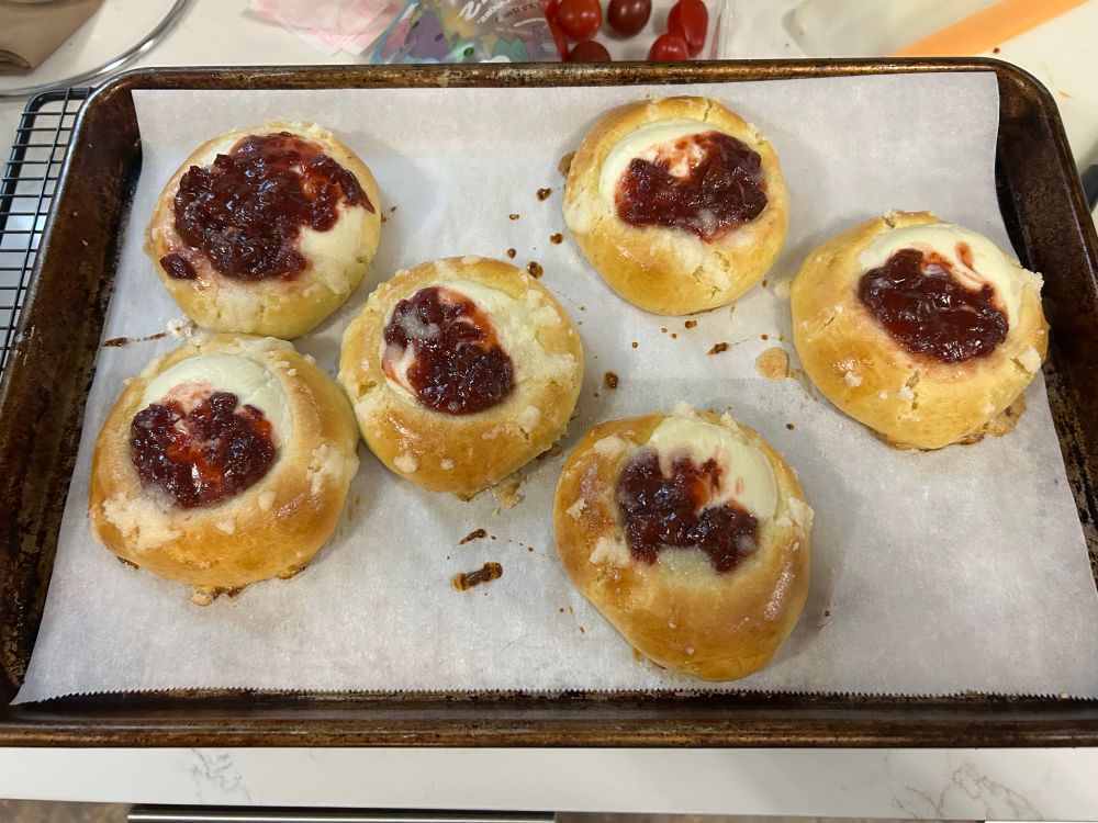Baking sheet holding six baked round pastries. They are light golden brown and filled with cheese and cherries. 