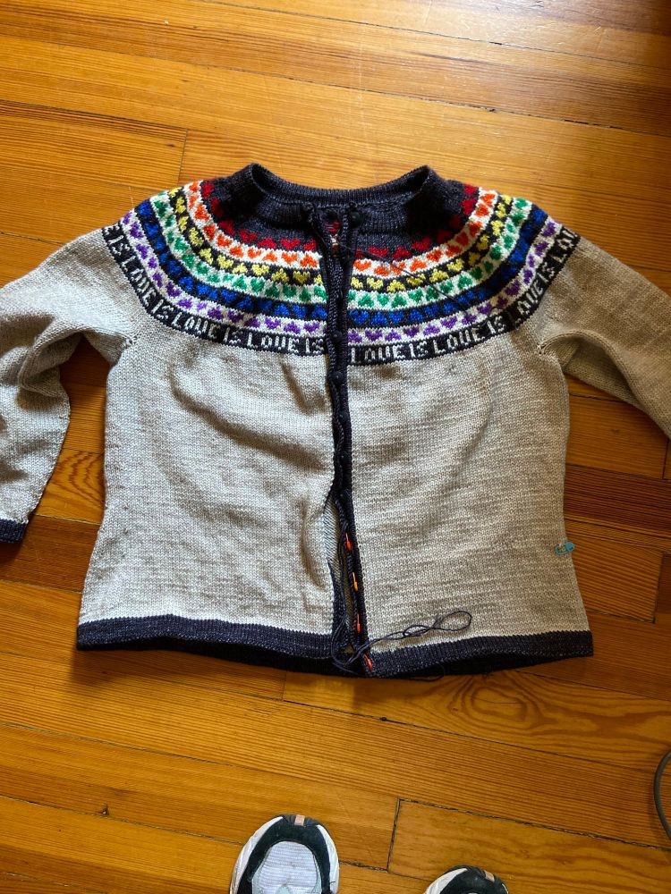 Handknit cardigan laid on the floor. It is light gray with black edging at the cuffs, hem, neckband and front bands. The yoke of the sweater is striped black and white. There are rainbow hearts on the stripes and text Love is Love. 