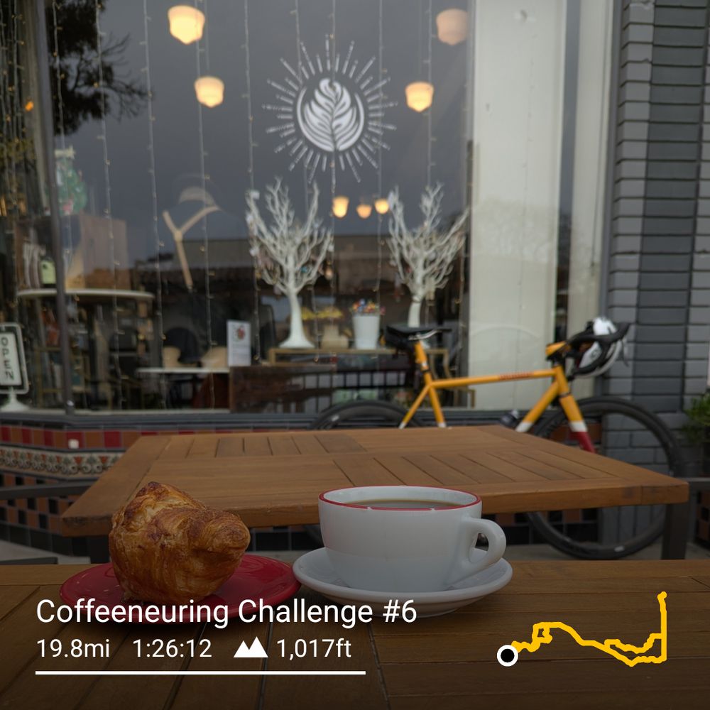 White coffee cup and saucer, next to a croissant on a red saucer. In the background is a yellow bicycle leaning against the storefront window that has a painted logo that looks like a latte.