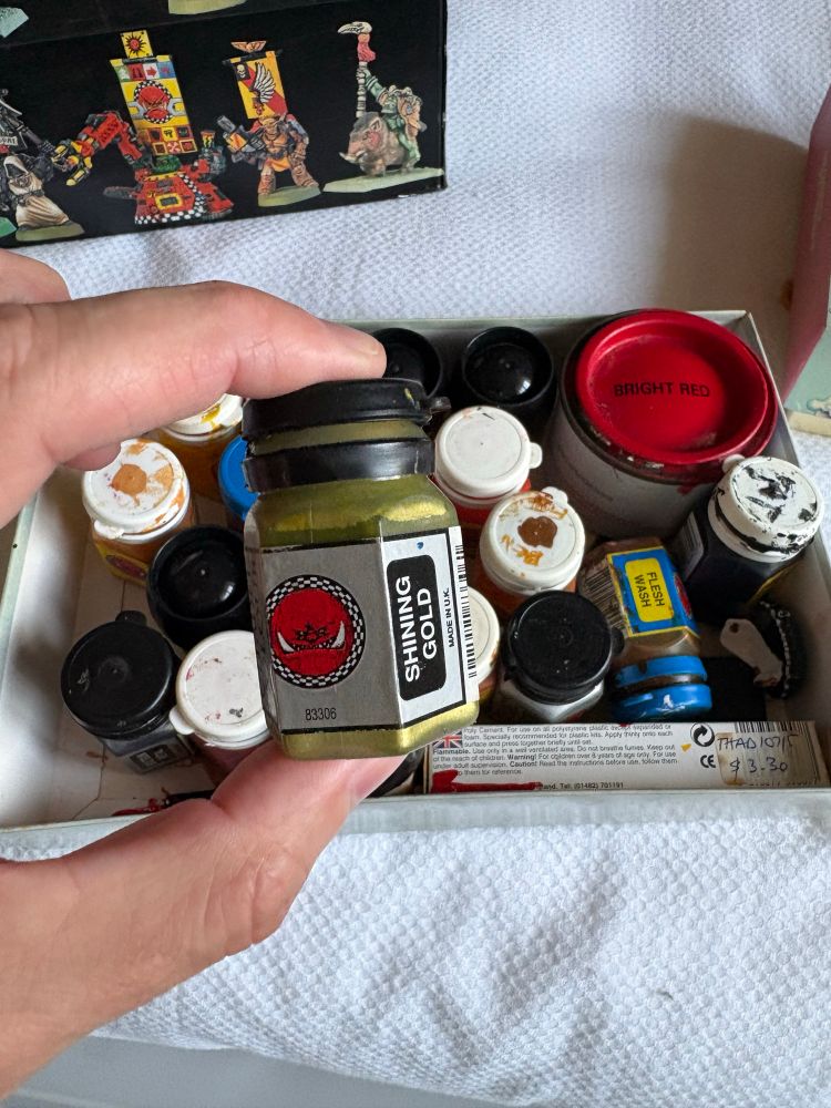 Old citadel paint pot “shining gold” being held in front of a box of old paints. 