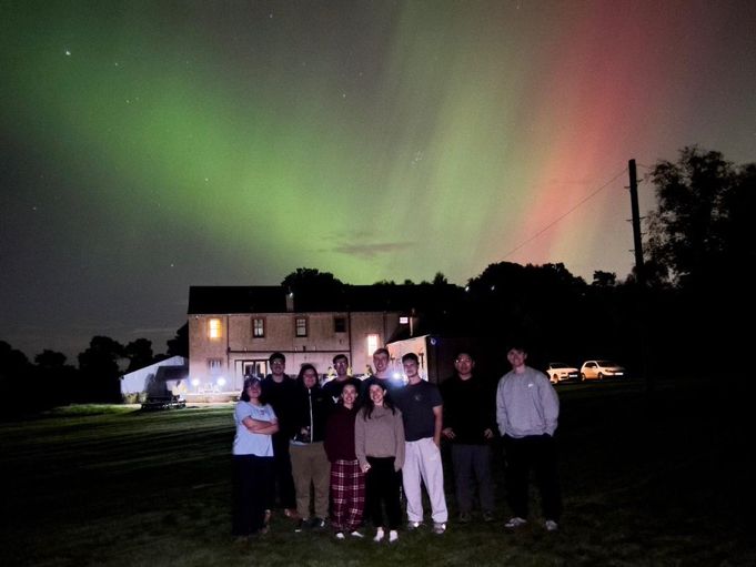The Lloyd-Jones Group (Summer 2024) standing in front of a house, the sky light with greens and reds of the Northern Lights