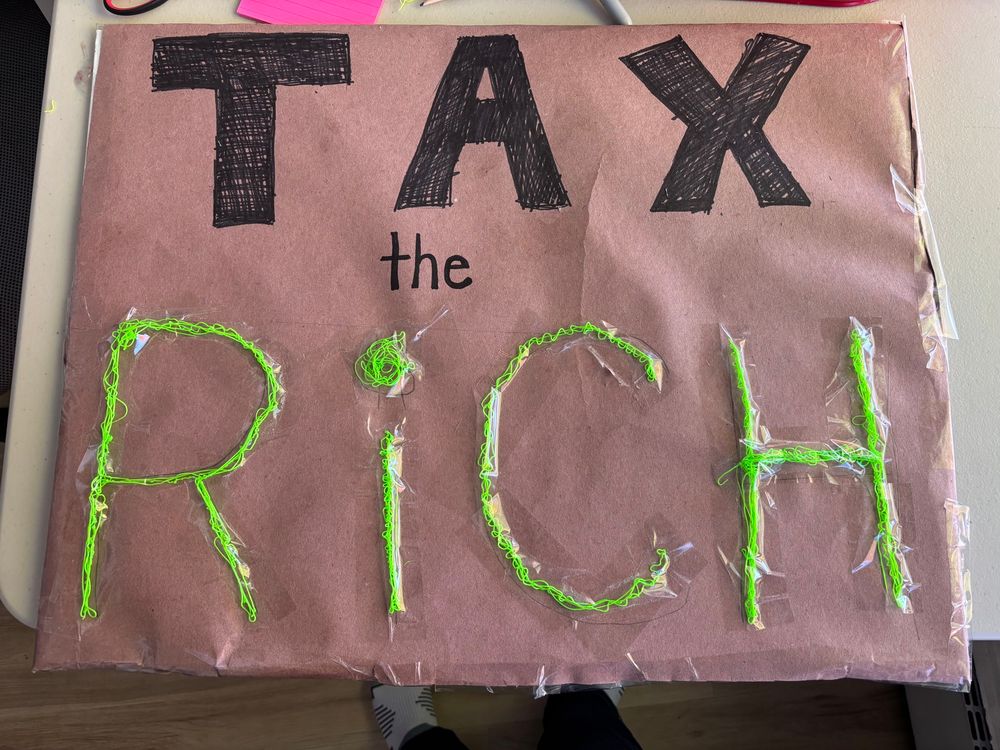 Pink poster with text. In black it says “tax the”, in green it says “rich” 