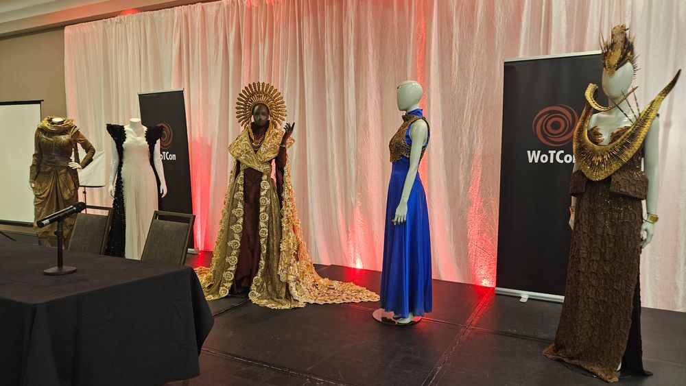This image showcases a fashion exhibition featuring stylized mannequins dressed in elaborate and culturally inspired outfits. The costumes appear to draw from African and possibly Afro-futuristic aesthetics, with rich textures, gold embellishments, and unique sculptural elements. The setting suggests a formal event with a backdrop featuring the logo "WoTCo" and promotional banners. 



