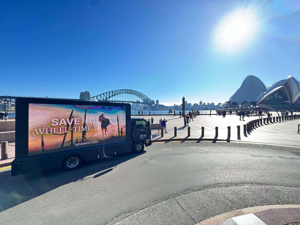 A brightly lit mobile billboard truck is parked on a paved area with a clear blue sky overhead. The truck's large side screen displays the text "SAVE THE WHEEL OF TIME" in large, bold letters, with a smaller "savewot.com" below it. The screen also features a visual of two figures walking away into a sunlit, hazy landscape. In the background, beyond the truck, are several iconic Sydney landmarks. To the left, part of the Sydney Harbour Bridge is visible. To the right, the distinct white sails of the Sydney Opera House stand out. Numerous people are walking around the paved area, and the sun is shining brightly, creating a glare in the upper right corner of the image. The overall impression is a sunny day in Sydney, with a clear focus on the promotional truck.