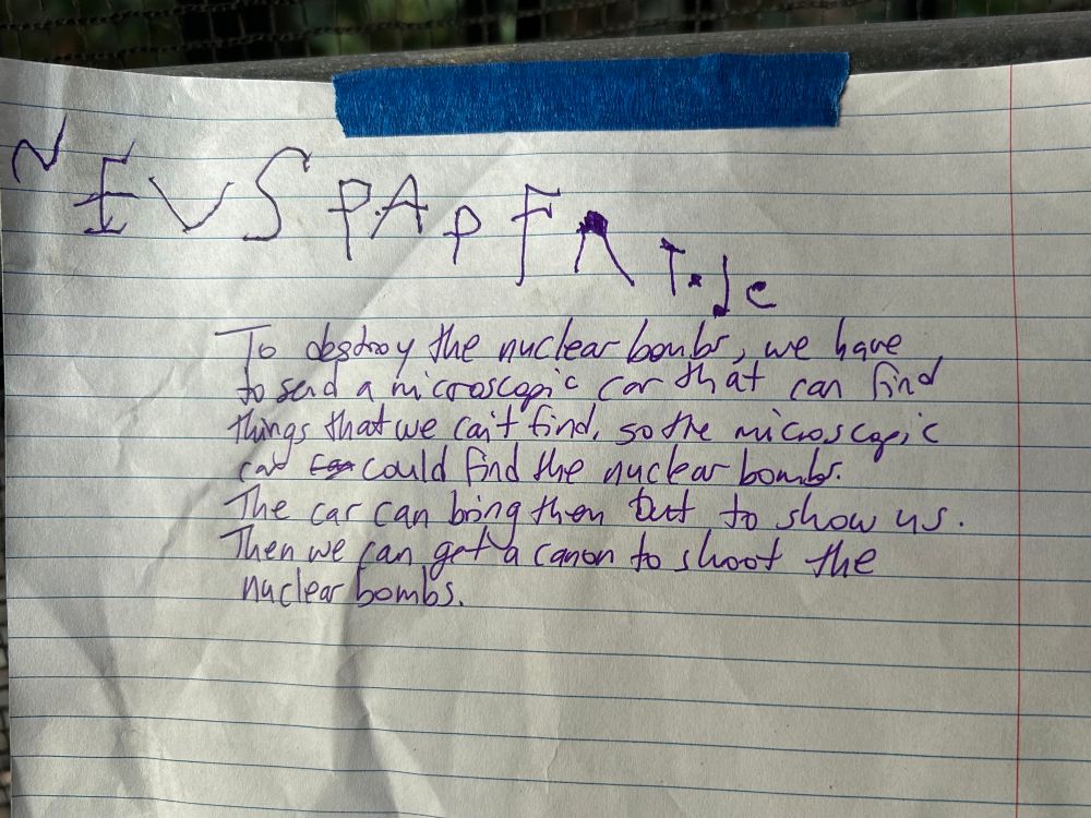 A page of lined paper taped to a fence. At the top, in a young child’s handwriting: NEUSPAP FR TODE. Underneath, in adult handwriting: To destroy the nuclear bombs, we have to send a microscopic car that can find things that we can’t find, so the microscopic car could find the nuclear bombs. The car can bring them out to show us. Then we can get a cannon to shoot the nuclear bombs. 