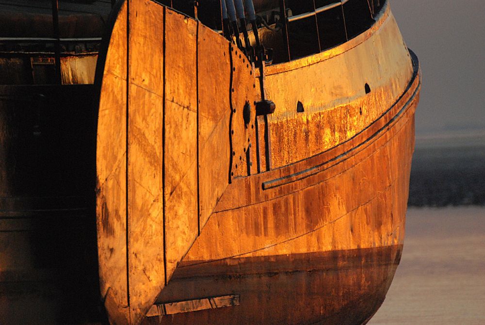 Op de foto staat een deel van de romp met het zwaard van een op het wad drooggevallen tjalk. Het schip wordt verlicht door een laagstaande ondergaande zon en lijkt in de weerspiegeling daarvan wel bedekt met een laagje bladgoud. Op het Groningerwad, Mothoek, september 2008.