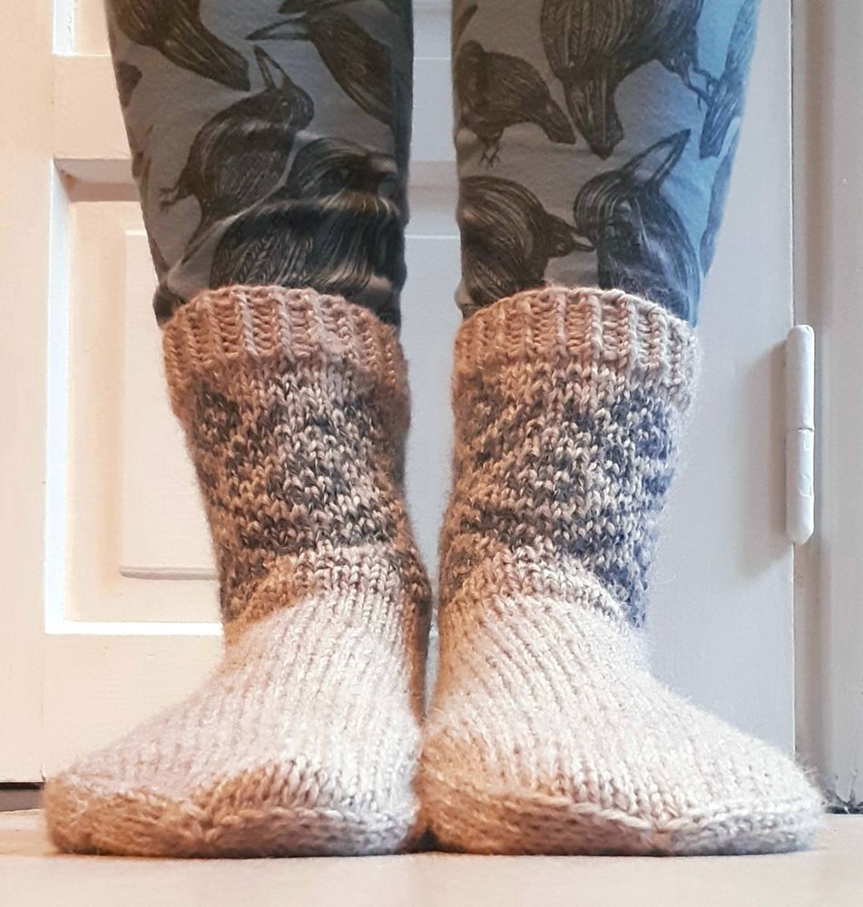 close-up of feet clad in linen coloured woolly socks with bluish pattern in stranded knitting around the ankles. above them are shown blue leggings with corvid illustrations in black.