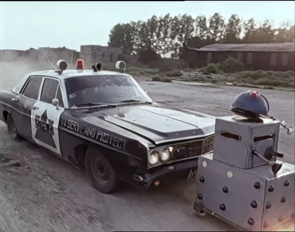 A still from the music video, Doctorin' The Tardis by The Timelords (a.k.a. The KLF).

A black and white American police car, looking the worse for wear, with “To Serve and Protect”, and The KLF logo (a ghetto blaster in front of a pyramid) on the side, pursues a D.I.Y. homemade "Dalek" on a dusty road.