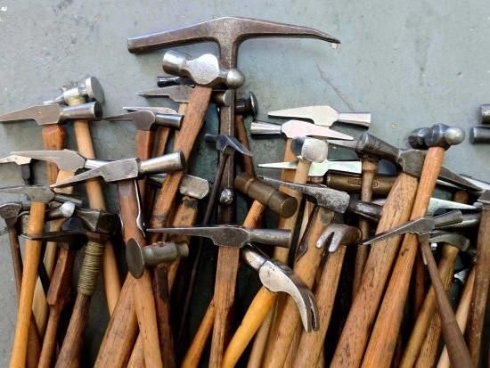 A pile of various hand-held hammers on a mid gray background, likely a workshop floor. The hammers are of different sizes and types.