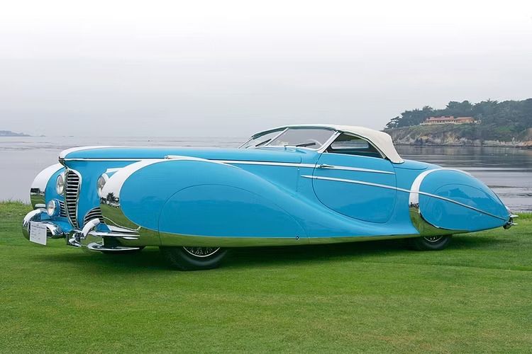 The photo shows a side-on view of a car, the 1949 Delahaye 175S Roadster.

Its paintwork is sky-blue with extensive chrome detailing. The swoopy, sinuous bodywork has covered wheel-arches (almost completely hiding the wheels) and is topped off with a white fold-down roof.

The car is parked on lush green grass that stands by the shore of a large body of water.

In the background, to the right of the photo, is a cliff with trees and a large house on top of it.