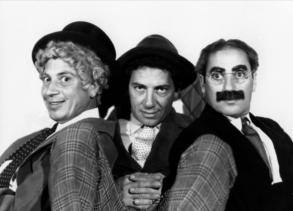 A black and white photograph of the Marx brothers. They are bunched close together and looking towards the camera. Harpo is on the left hand side of the image, with Chico in the middle and Groucho on the right.
