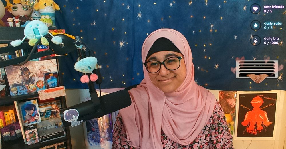 a photo of sahar smiling. they are wearing a light pink hijab and a black dress decorated with pink flowers. around and behind them are various pieces of fan merch and art prints.