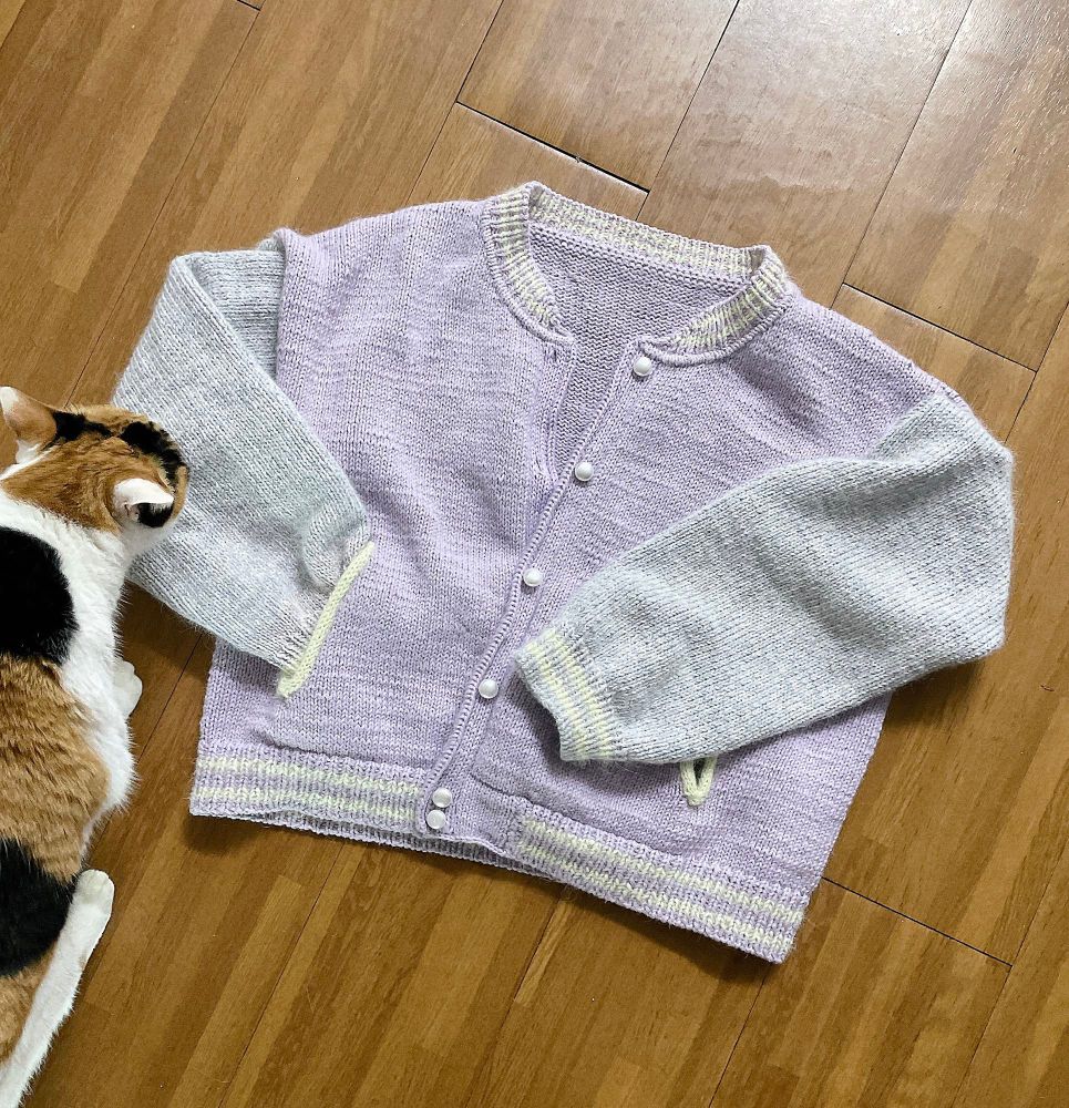 연보라색 몸판과 회색 소매가 달린 손뜨개 야구점퍼. 그리고 왼쪽엔 삼색 고양이가 냄새를 맡고 있다.