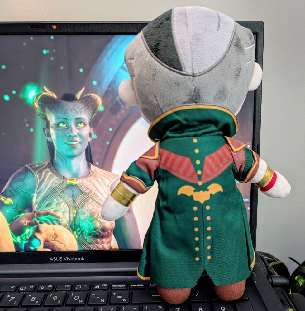 The back of a gorgeous handmade plush of the character Emmrich from Dragon Age: The Veilguard. He's propped up against an open laptop with a screenshot of Ruya "Rook" Laidir looking at him in wonder. 
