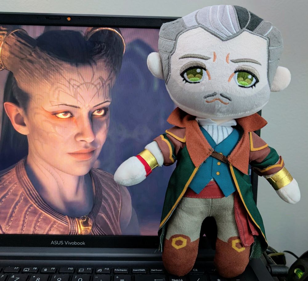 A gorgeous handmade plush of the character Emmrich from Dragon Age: The Veilguard. He's propped up against an open laptop with a screenshot of Ruya "Rook" Laidir giving him bedroom eyes. 