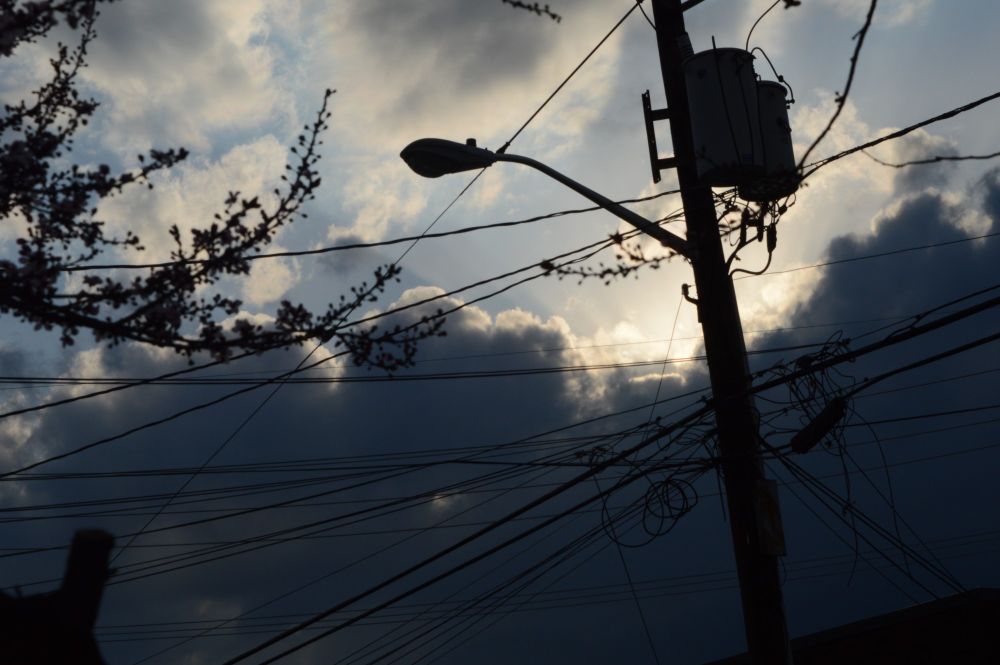 A picture of the sun peeking out of the dark clouds, with telephone wires and a street lamp in the forefront.