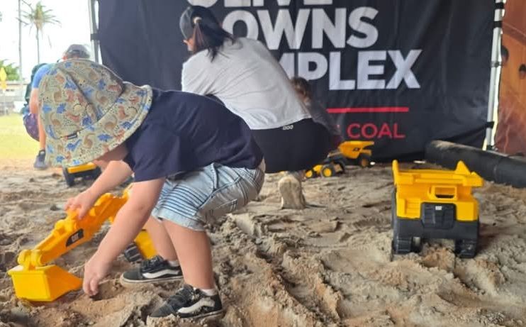 child crouched playing with a yellow digger in a sandpit. at back is a banner with the word coal. rest of the banner is hidden behind a crouching adult