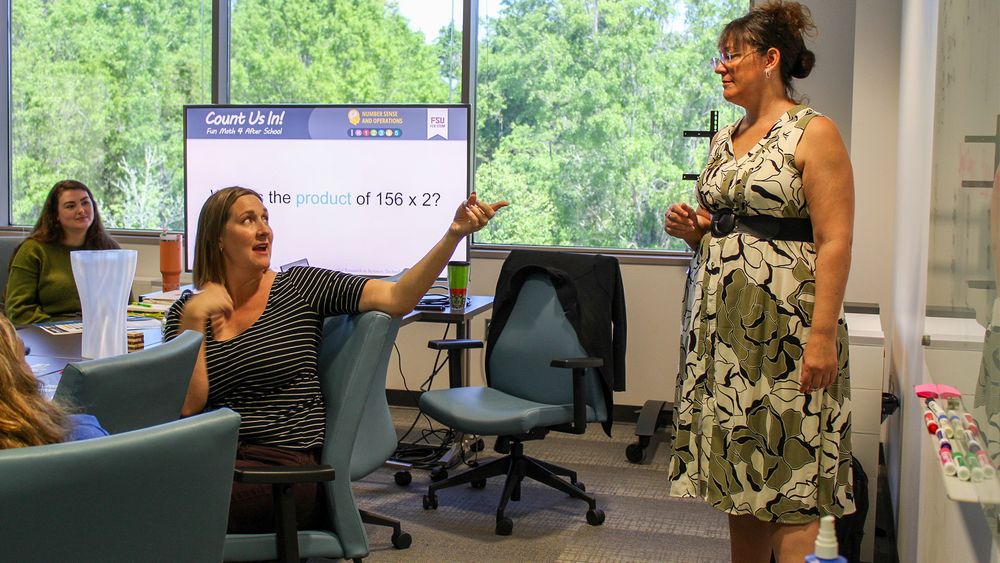 A discussion between two people. One is seated at a conference table turned in her chair talking to another person who is standing.