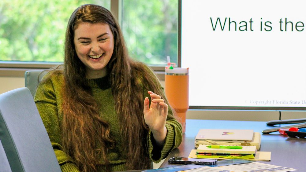 Woman sitting at a table laughing. Behind her is a large TV screen with part of a phrase "What is the"