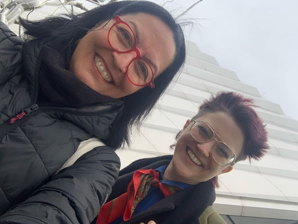 on the left is a Mexican woman (Ana) with shoulder-length hair. She is wearing a black puffer coat and red glasses and has an inviting smile. Her left arm is out because she's taking the selfie of us. I am on the right. I'm a nonbinary white person wearing glasses with gold rim and a silver septum piercing. I have a purple-red mullet. I am wearing a grey wool coat, my great-grandmother's red scarf, and a blue coverall. I'm looking so thrilled with squinting eyes because I finally got to meet Ana in person. In the background is the beautiful ripple texture of the MSU Broad Art Museum. The photo is to a diagonal angle. 