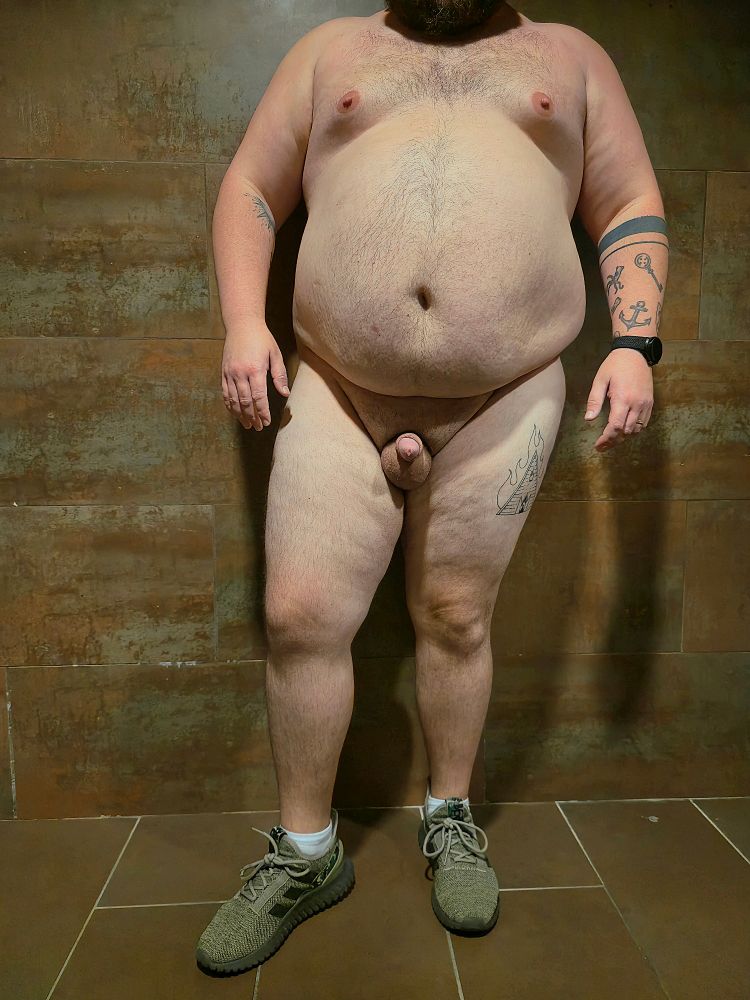 Fat man standing nude in the public bathroom posing to show off his new shoes .