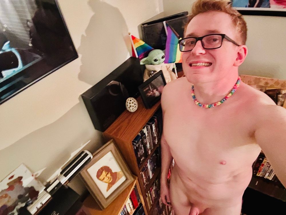 Selfie. I’m in a corner of my living room surrounded by movie shelves. Naked except for my glasses & rainbow puka necklace. New short haircut letting my natural ginger hair return. A toy Grogu is waving Pride flags on the shelf. 