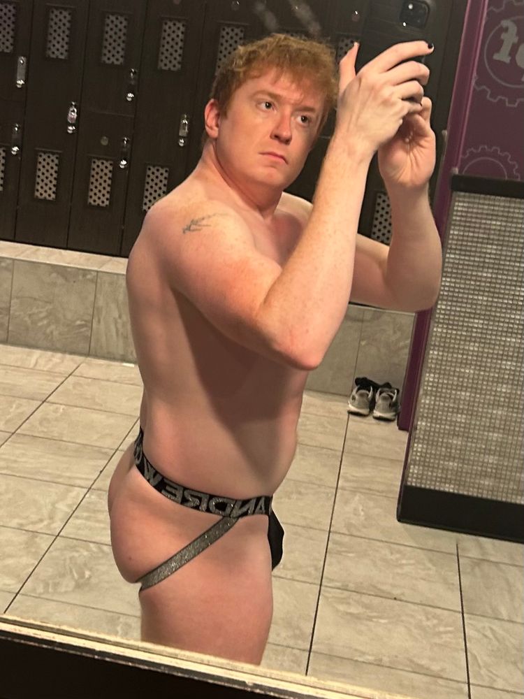 Locker room selfie in a black Andrew Christian jock with silver straps. Side view to show cheek & slight cage outline. 