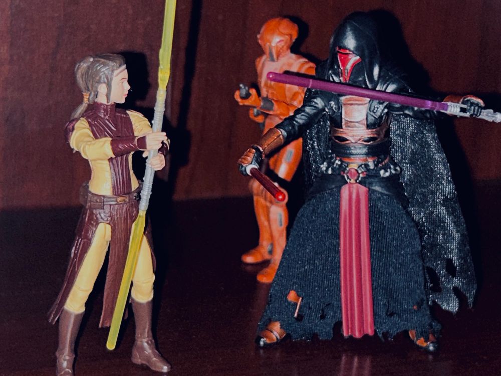 Bastila Shan, HK-47, & Darth Revan action figures. Bastila is confronting Revan & his droid. Bastila holds her double-bladed yellow lightsaber. Revan holds his red lightsaber forward and his purple lightsaber to the side. HK-47 aims his rifle at Bastila. 