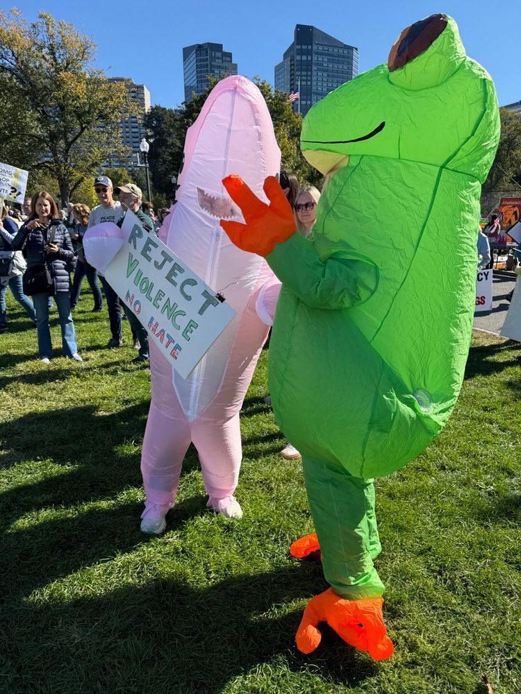 Frog and shark inflatable costumes at the No Kings rally. The shark holds a sign that says REJECT VIOLENCE NO HATE