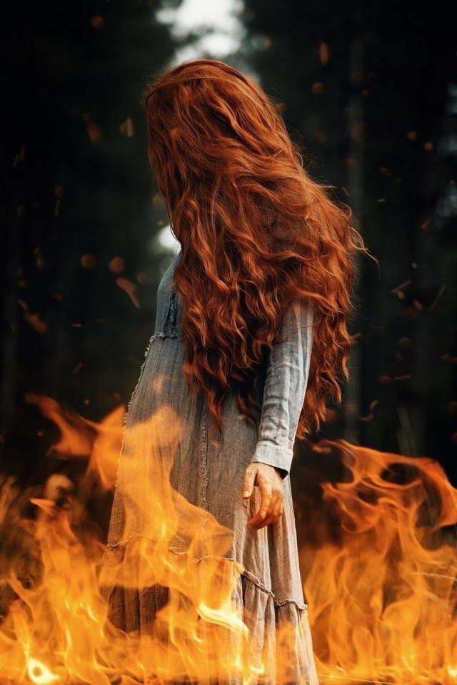 A redhead standing in fire with a forest backdrop. 