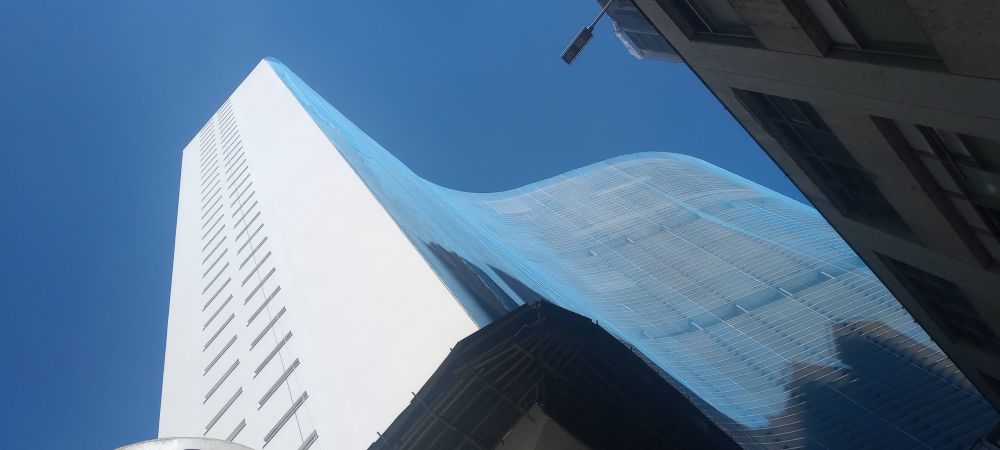 Vista de baixo para cima do Edifício Copan, ao fundo um céu azul combinando com o azul da tela de segurança que cobre a faixada sinuosa do prédio 